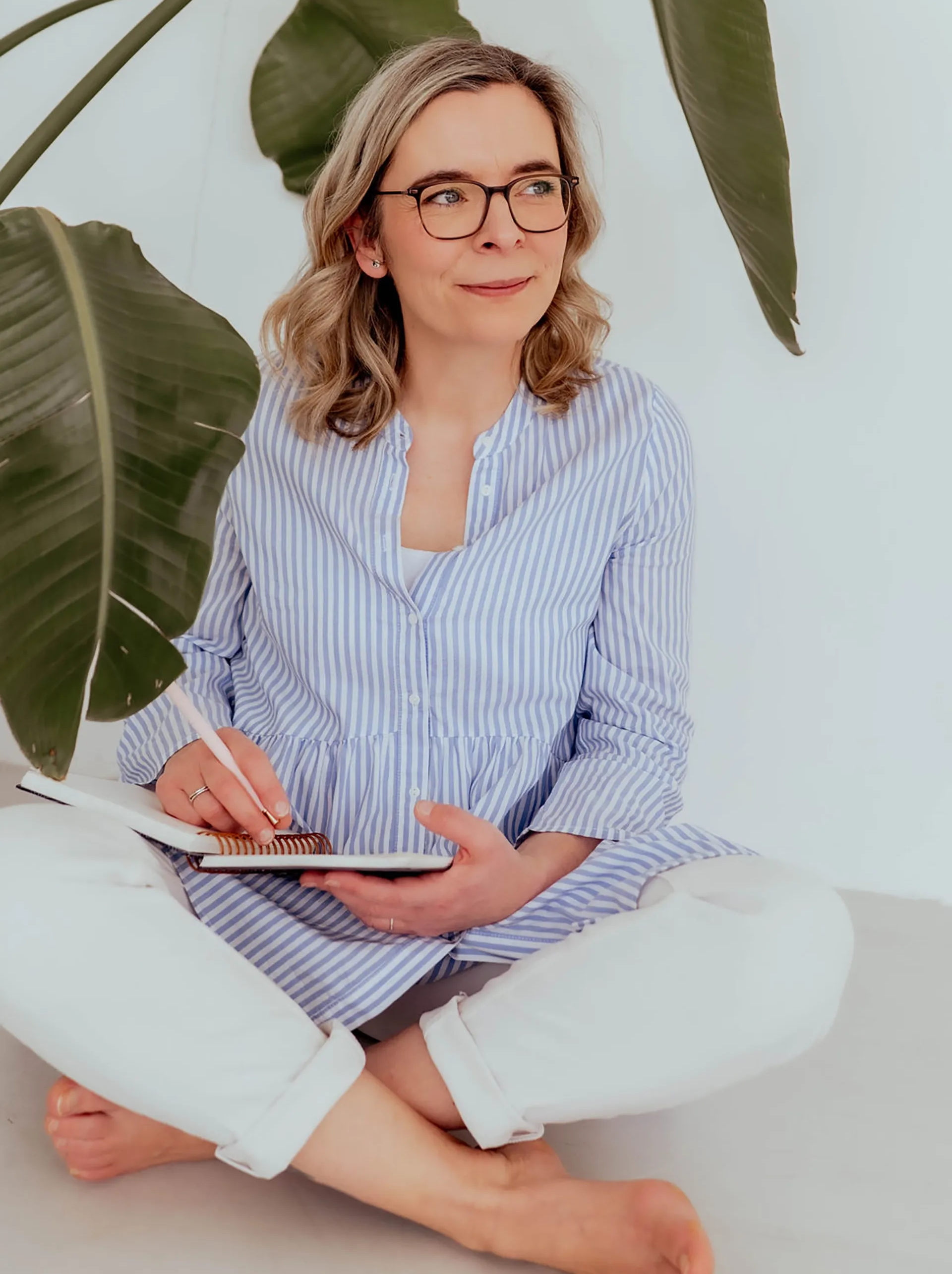 Eine Frau mit Brille sitzt im Schneidersitz neben großen grünen Blättern und schreibt in ein Notizbuch.