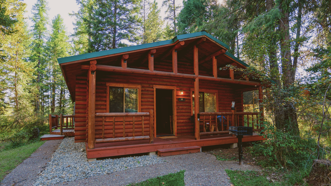 Wooden cabin with a porch in a forest, surrounded by tall trees and gravel path
