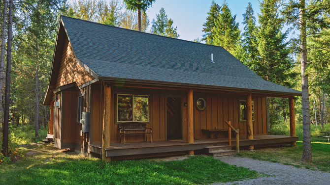 Rustic wooden cabin with dark roof in a pine forest, with a porch and green grass around it