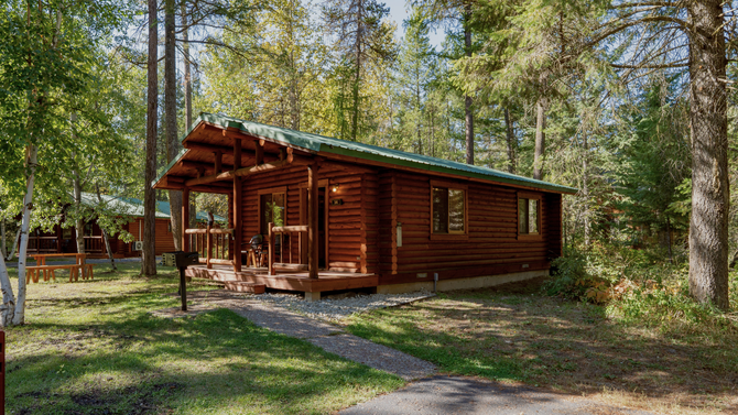 Wooden cabin in a forest clearing, with a small porch and trees surrounding it.