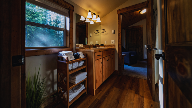 Cozy wooden bathroom with a sink, mirror lights, window, and a blue rug leading to a shower.
