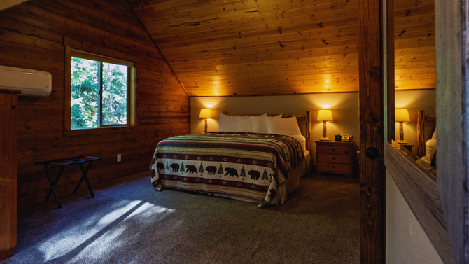 Cozy wooden cabin bedroom with a large bed, warm lamps, and a window overlooking trees.