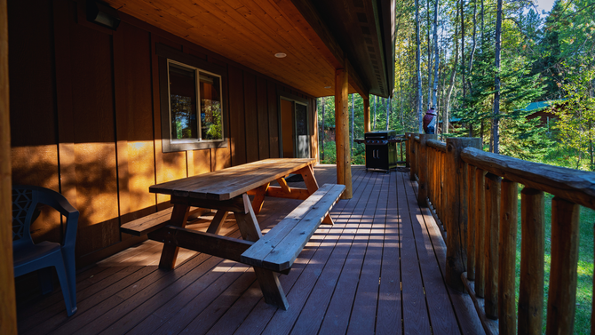 Wooden cabin porch with picnic table, railing, and sunlit forest view beyond.