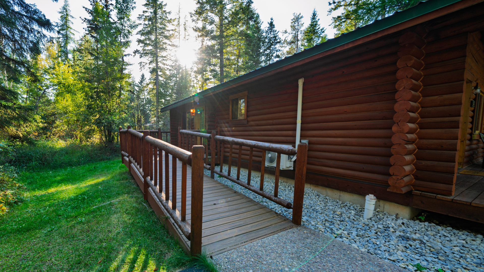 Wooden cabin porch with railing beside a gravel path, surrounded by green trees in warm sunlight
