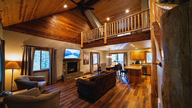 Cozy log cabin living room with wood-paneled walls, stone fireplace, loft, and kitchen area under warm lighting