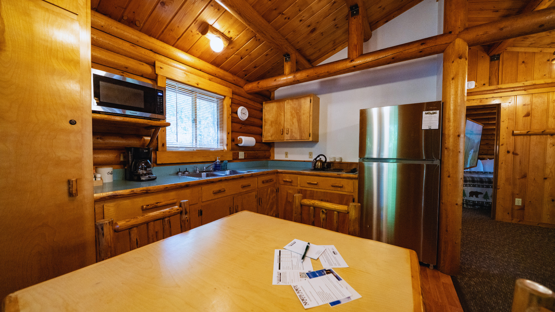 Wooden cabin kitchen with pine ceiling, stainless fridge, and papers on a table.