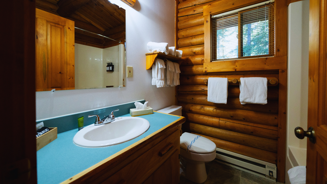Small rustic bathroom with wood log walls, turquoise countertop sink, toilet, mirror, and towels by a window.