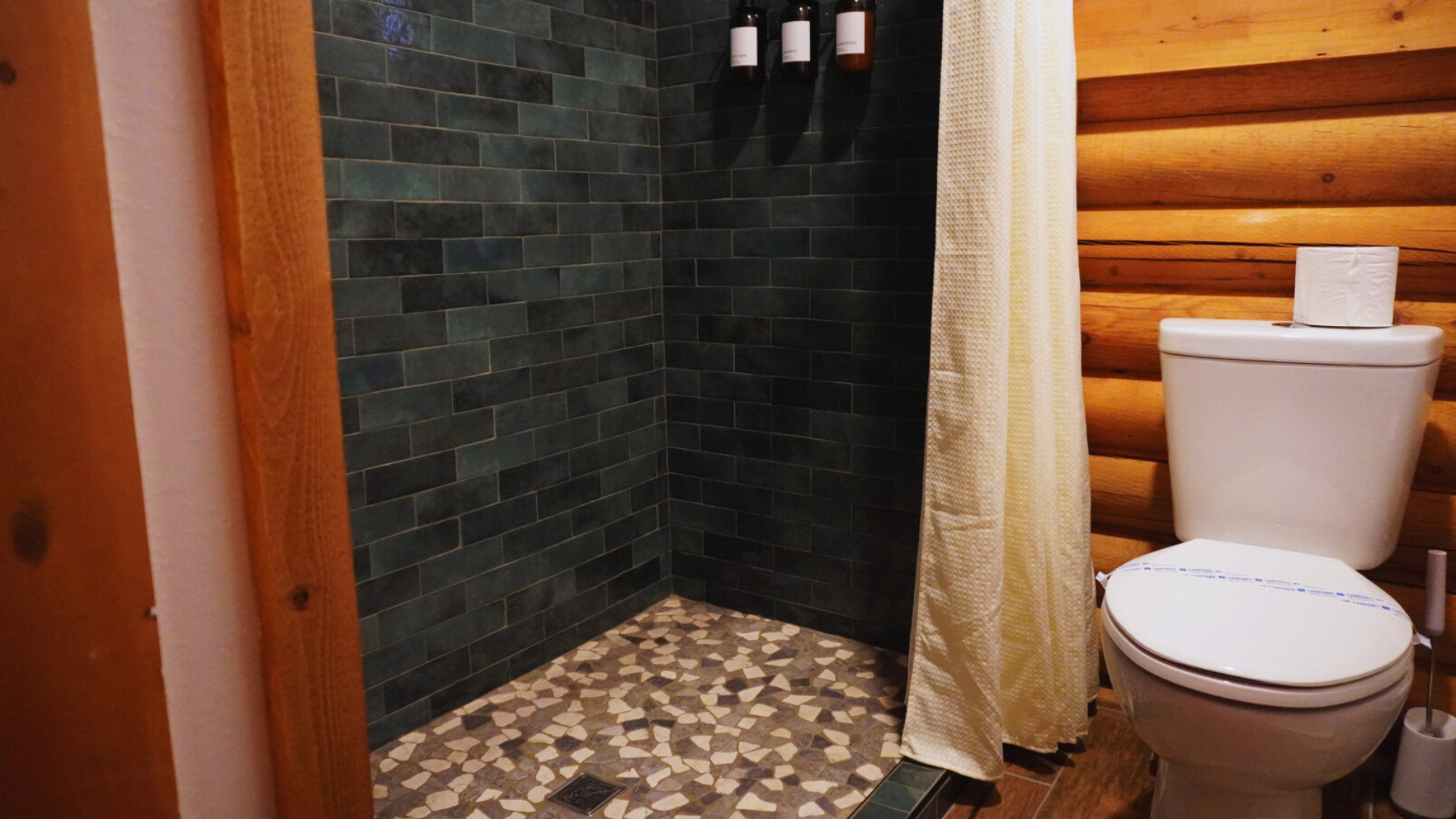 Rustic bathroom with log walls, tiled shower, and white toilet beside a curtain