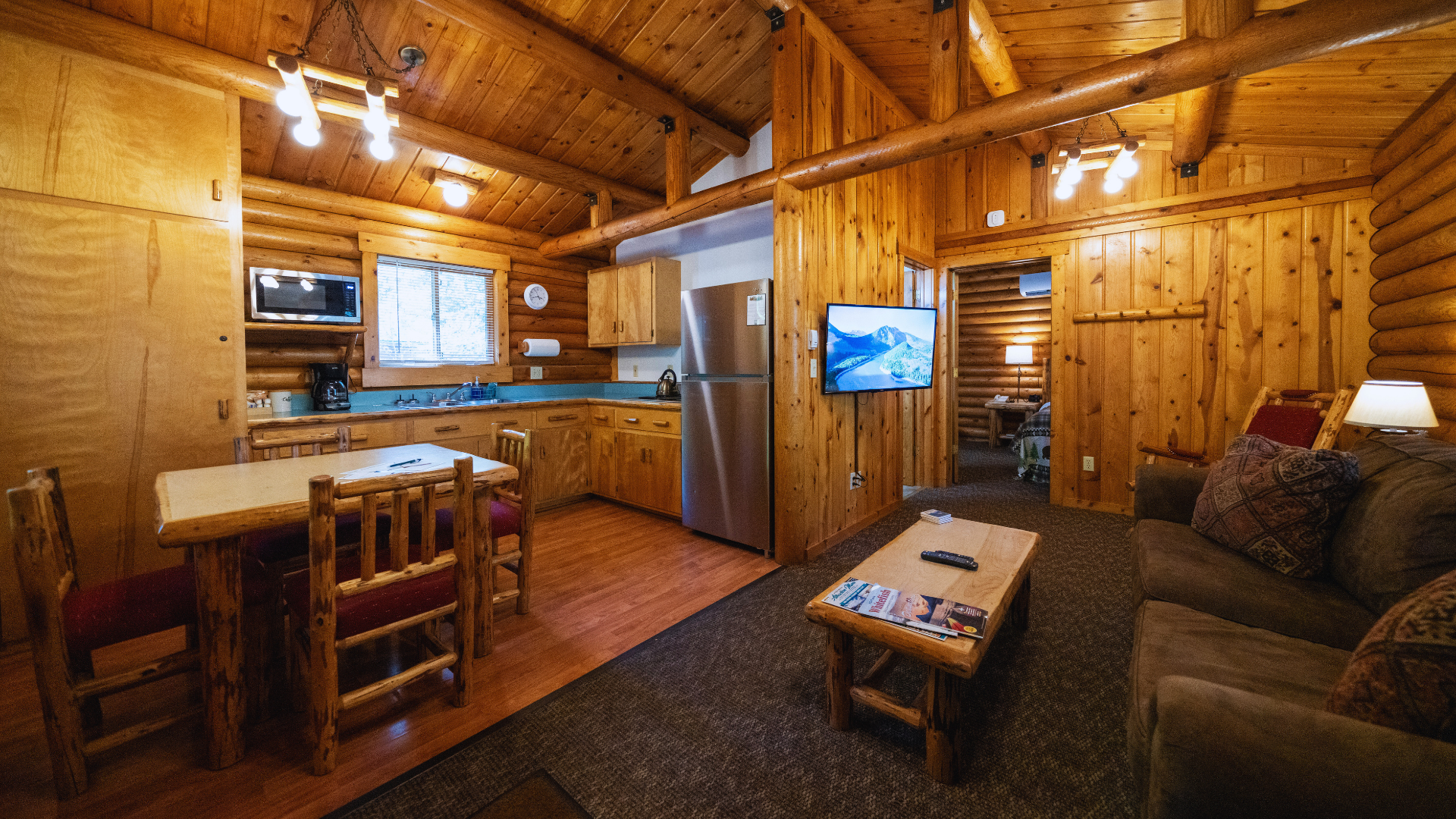 Cozy wooden cabin living room with kitchen, sofa, coffee table, and TV on warm wood-paneled walls