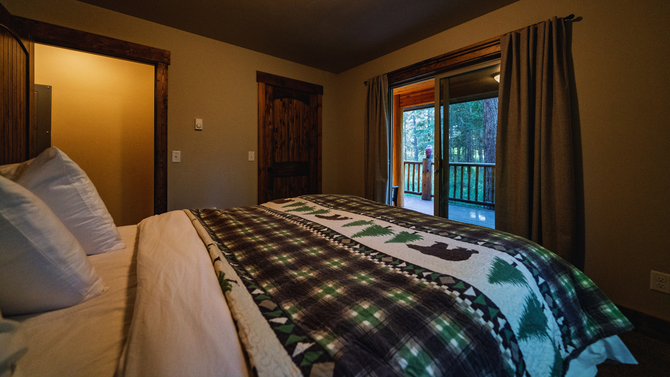 Cozy bedroom with a made bed, plaid blanket, warm lamp light, and sliding glass doors to a balcony