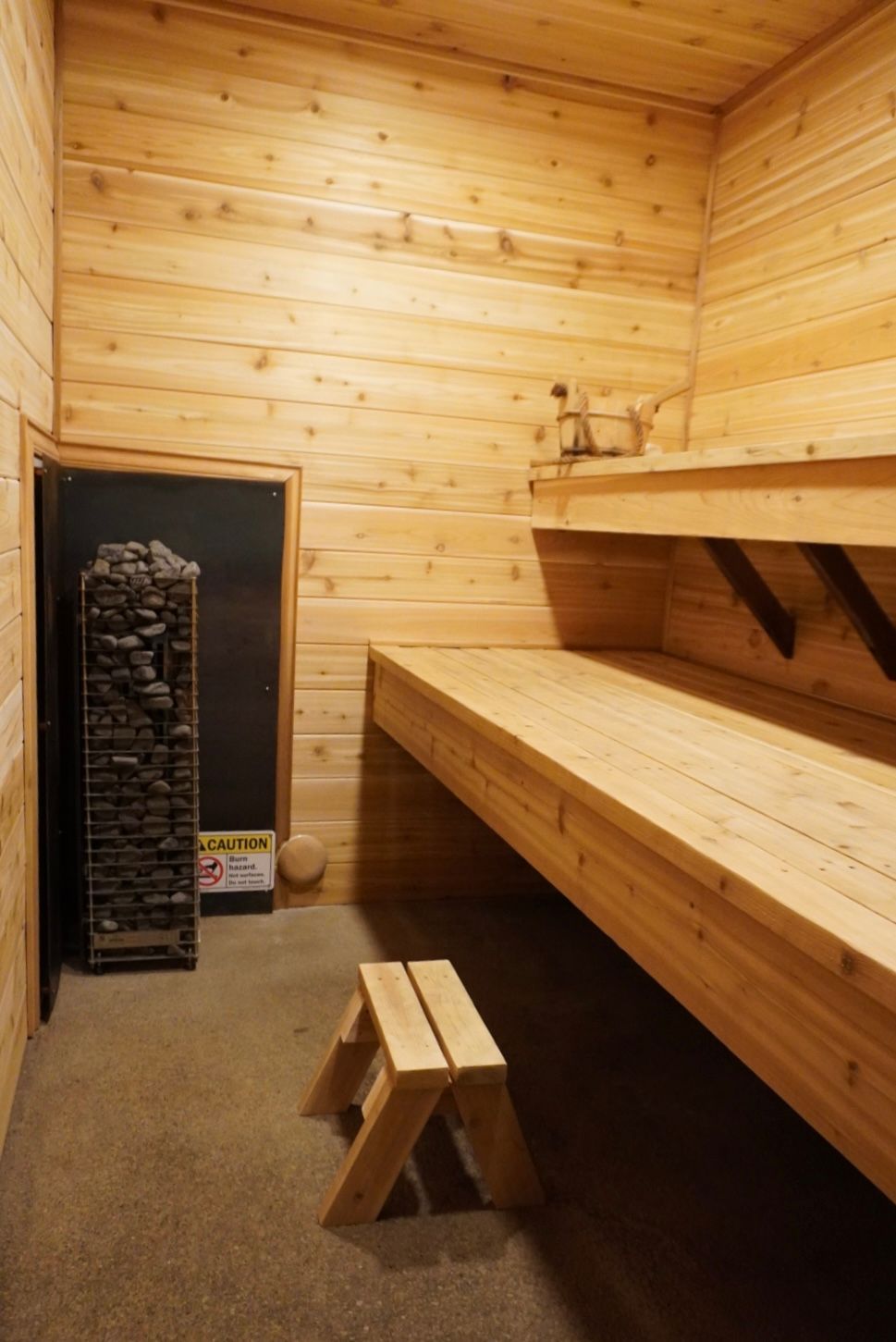 Wood-paneled sauna interior with wooden benches, a heater, and a small stool in the center