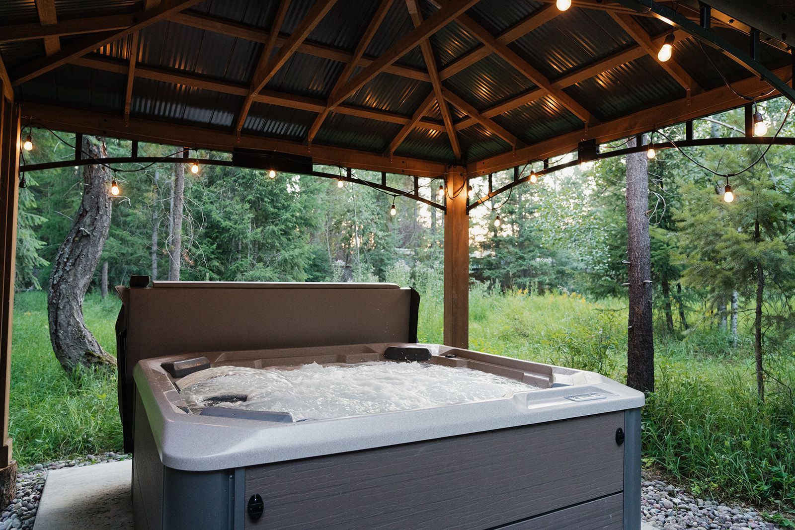 Hot tub under a wooden gazebo in a wooded backyard, with forest greenery surrounding it
