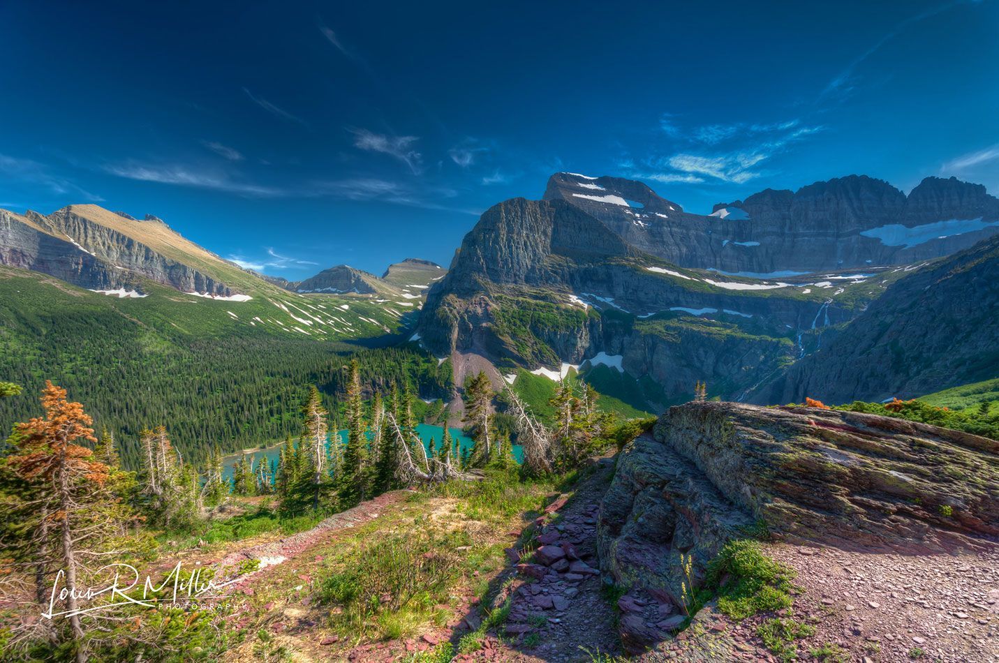 Mountain landscape with a turquoise lake, green forest, and snow-capped peaks under a blue sky.