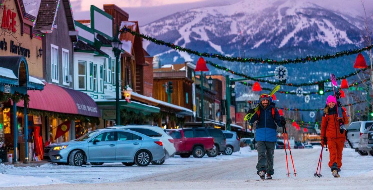 People with skis walk on a snowy street in a mountain town, festive lights overhead, snowy mountains in the background.