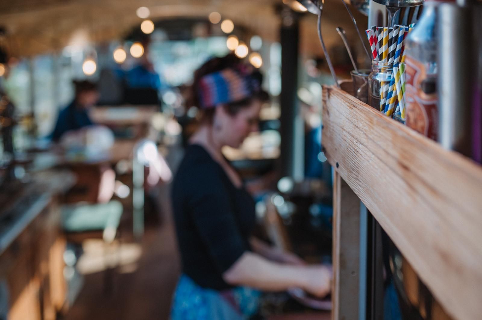 Vrouw werkt in een café, met een haarband en schort aan, met wazige klanten en verlichting.