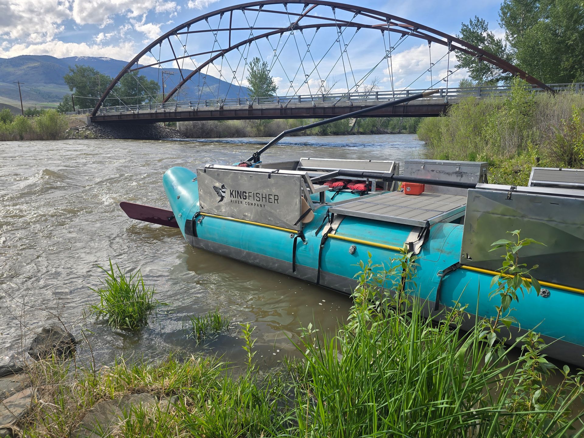 Blue raft in shallow river beneath a metal arch bridge, with mountains in the background.