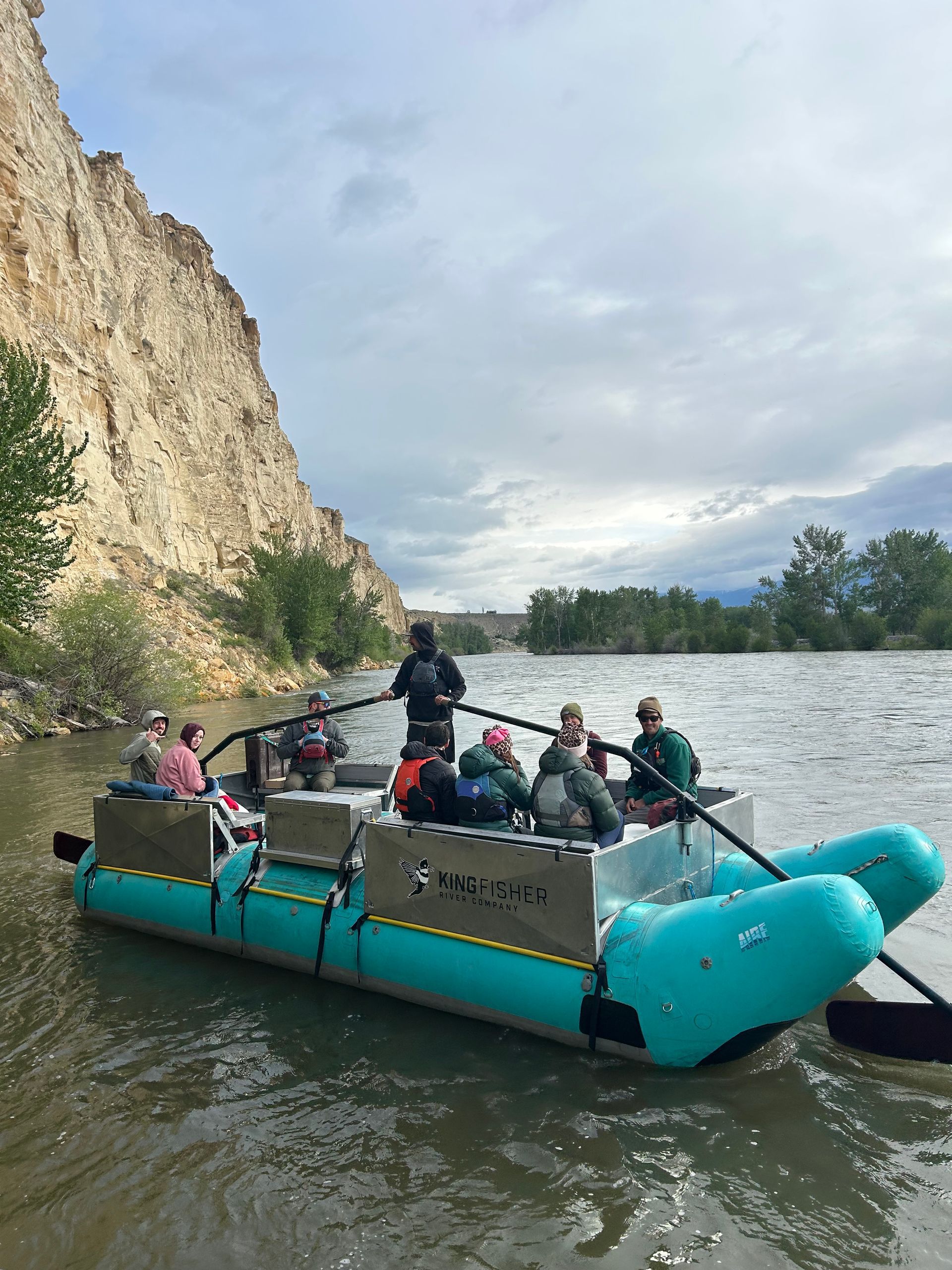 People on a turquoise pontoon boat on a river beside a rocky cliff and trees under cloudy skies