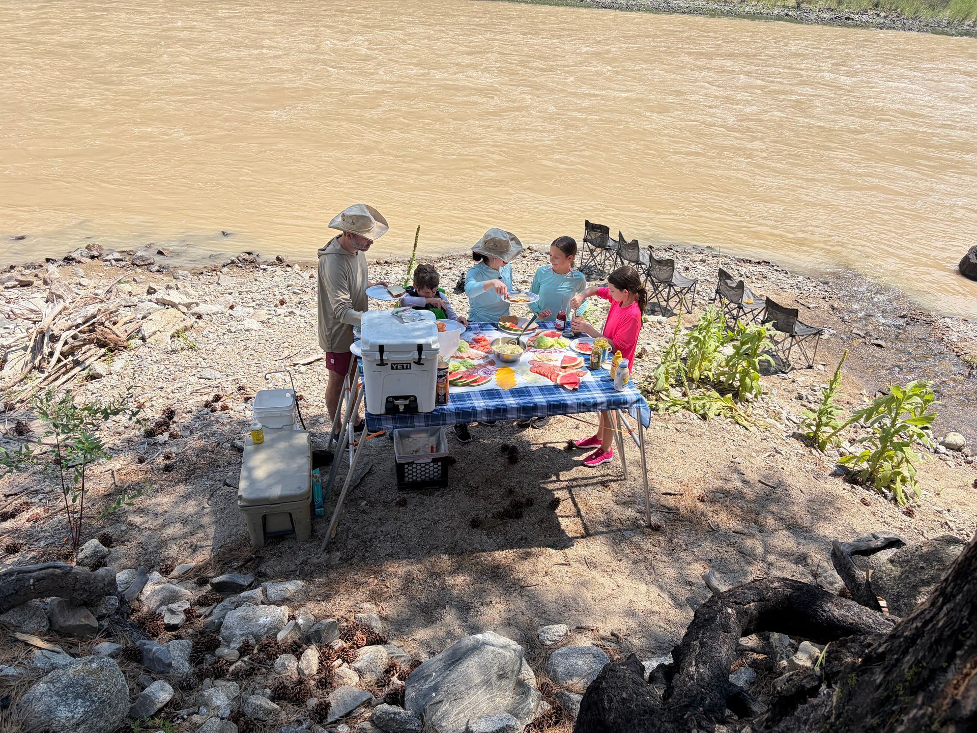 People gathered around a picnic table on a rocky shore beside calm water.