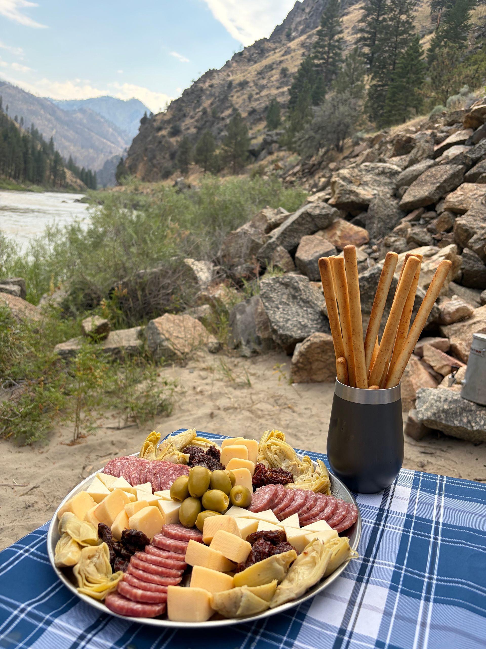 Charcuterie board and breadsticks on a blue blanket beside a river in a rocky mountain valley