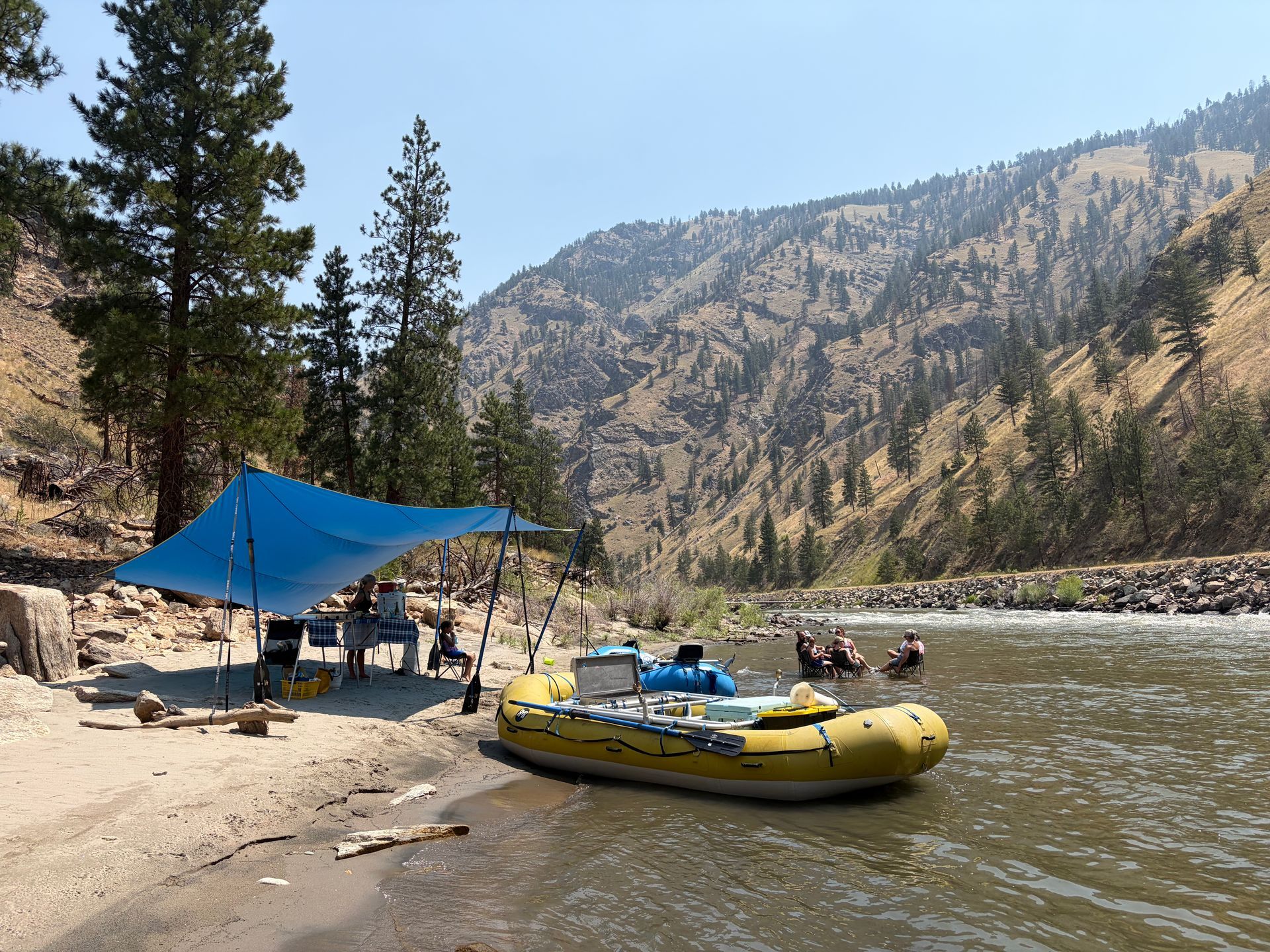 Rafting camp by a river with yellow rafts, blue canopy, and pine-covered mountains
