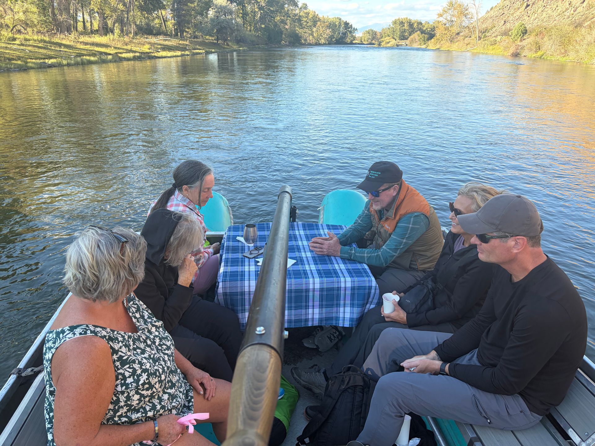 Six people sitting at a river picnic table, chatting beside calm water and trees.