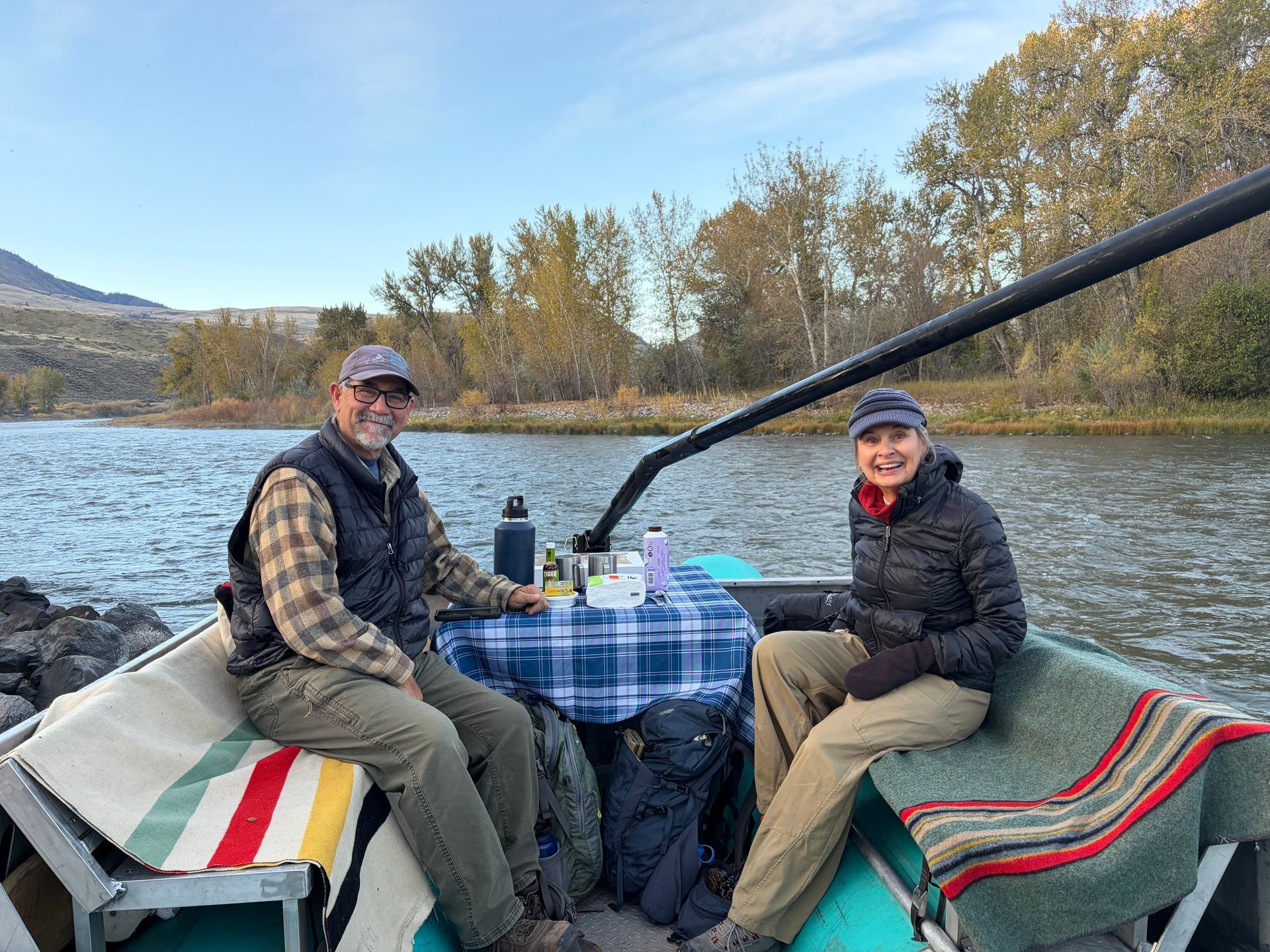 Two people seated in a boat on a river, with blankets, a blue checkered tablecloth, and autumn trees in the background