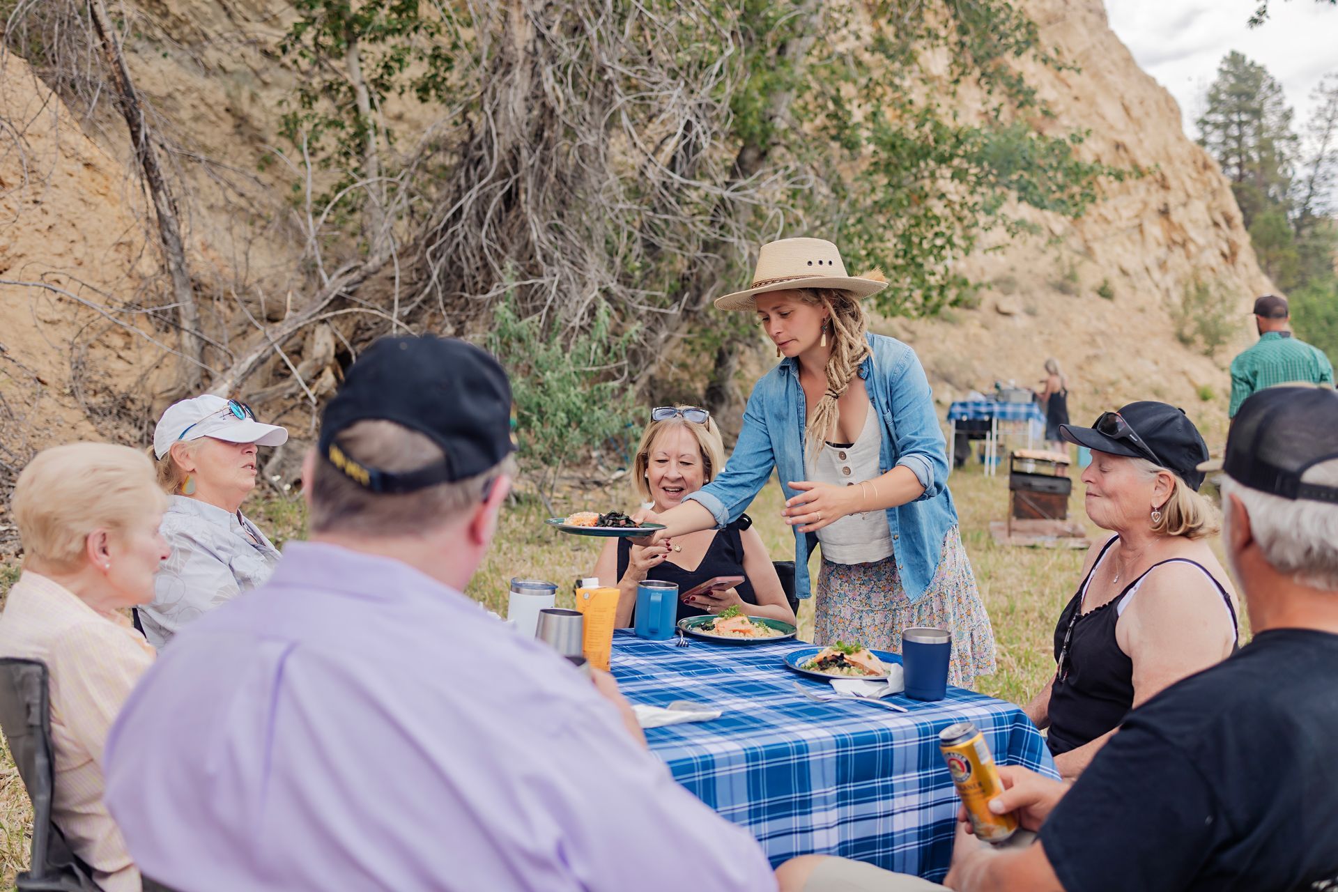 Group gathered around a blue-checkered picnic table at an outdoor cliffside meal, with a woman serving food.