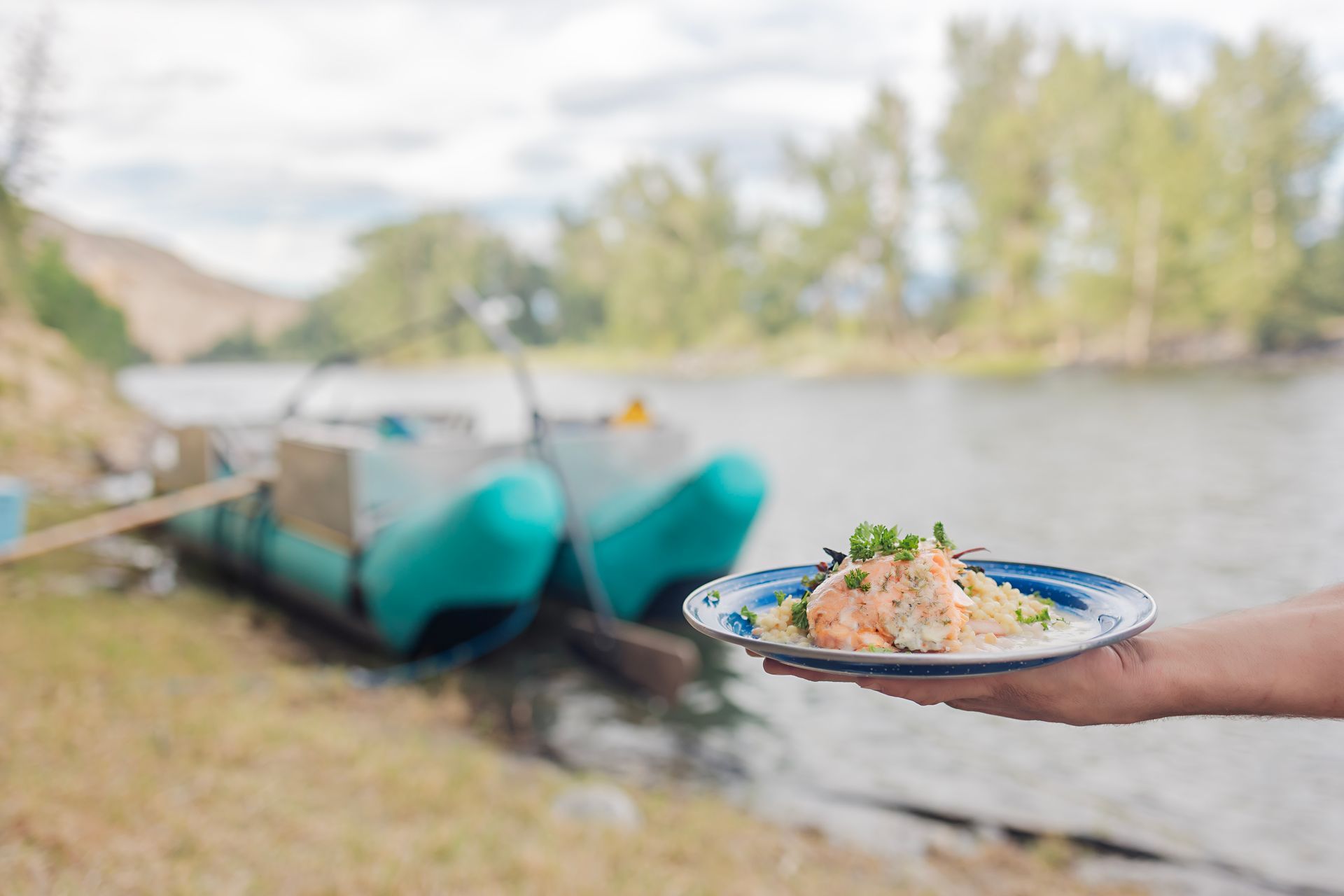 Hand holding a plate of food by turquoise boats on a calm riverside.
