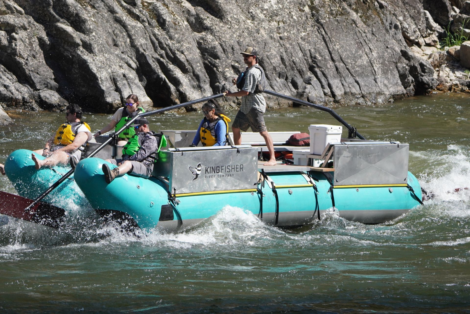 Whitewater rafting on a rocky river, with several people in a teal raft splashing through rapids.