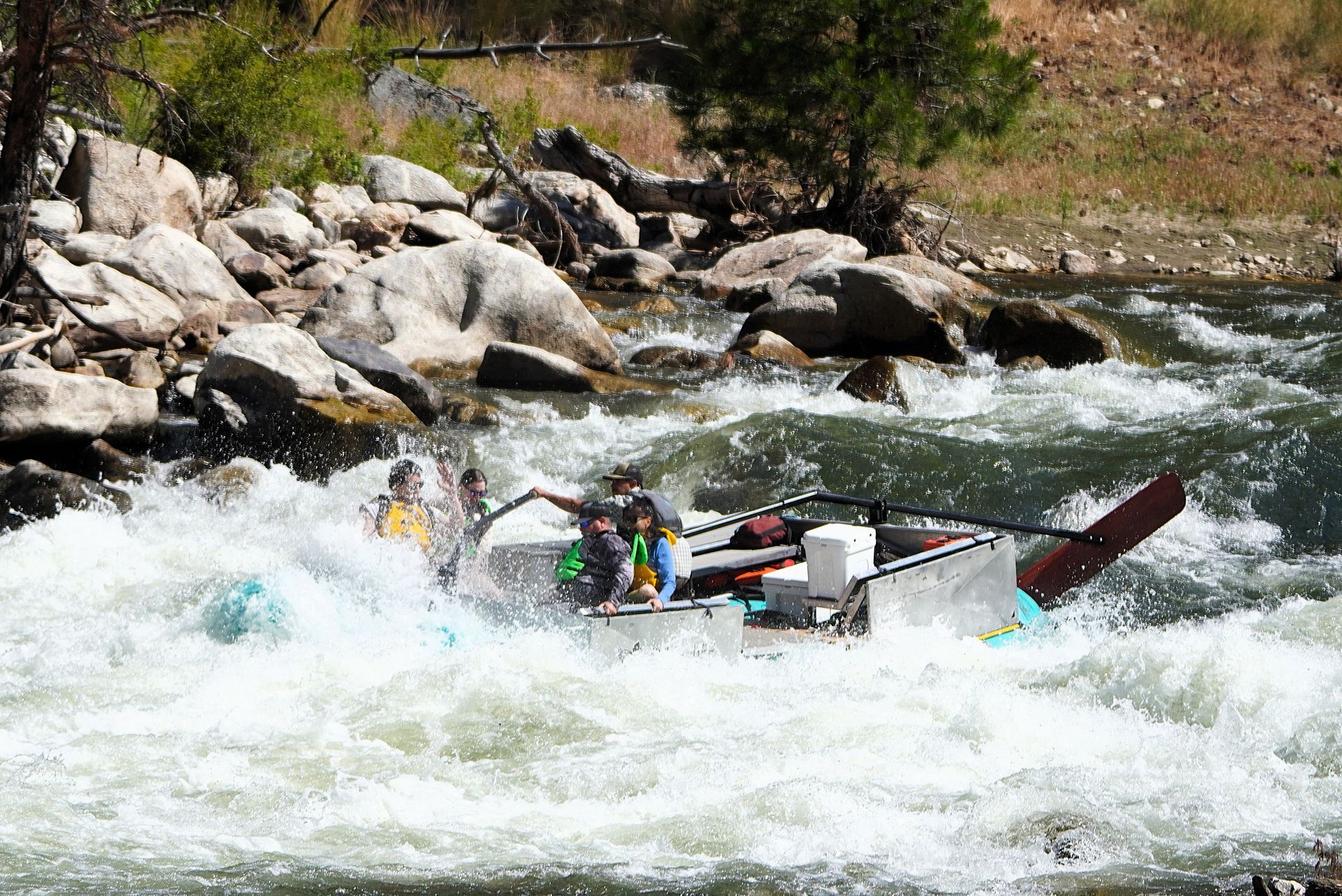 Rafting team navigating whitewater rapids beside rocky riverbank, splashing through rough water.