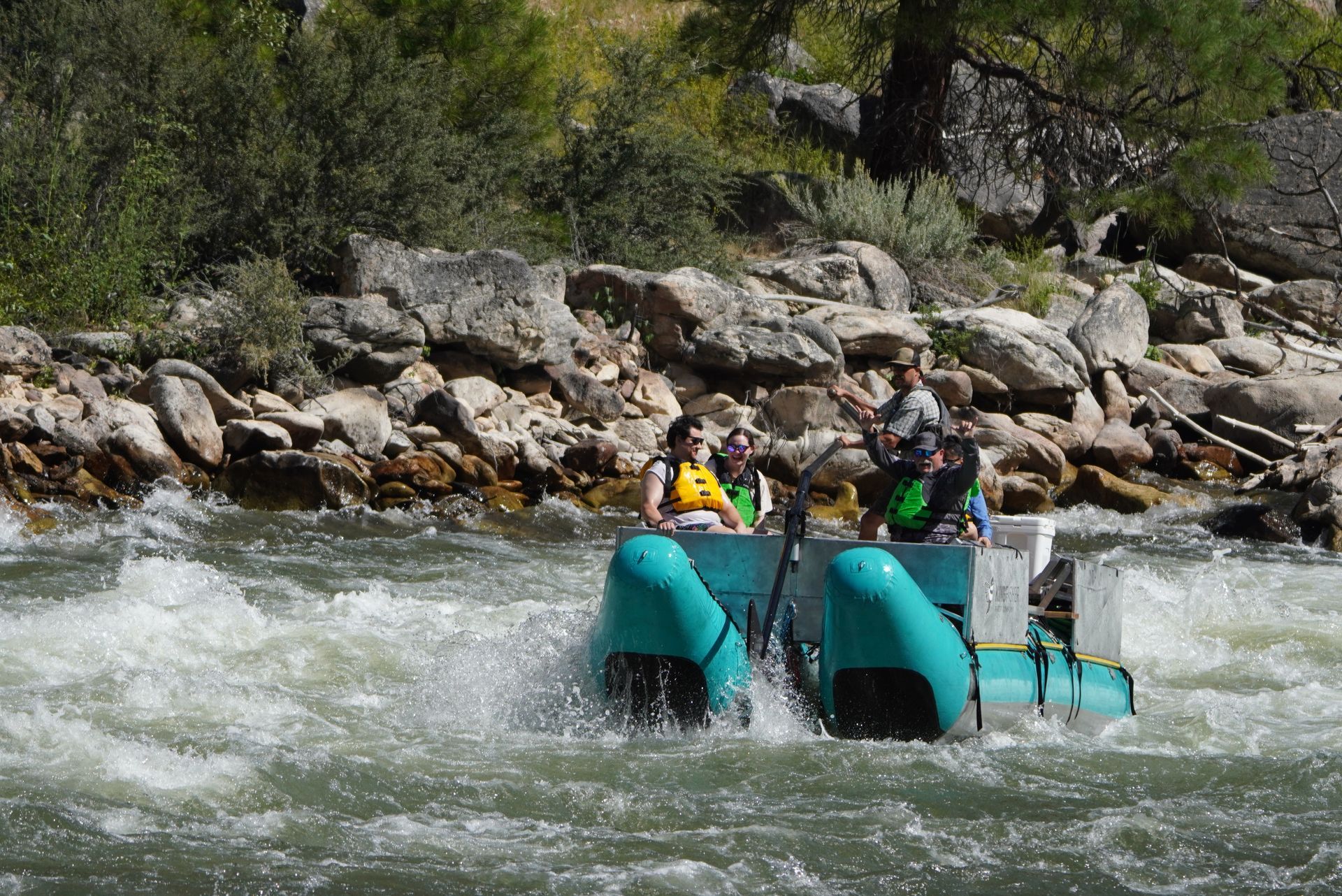 Raft with two people speeding through whitewater beside a rocky riverbank