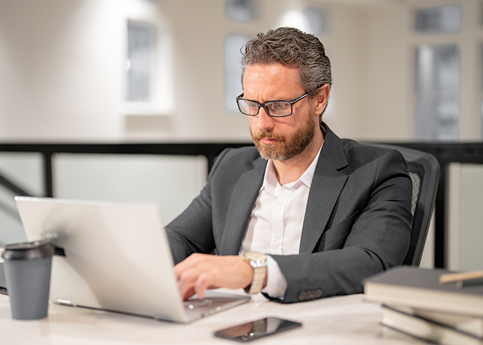 Un hombre con traje y gafas está sentado en un escritorio utilizando una computadora portátil.