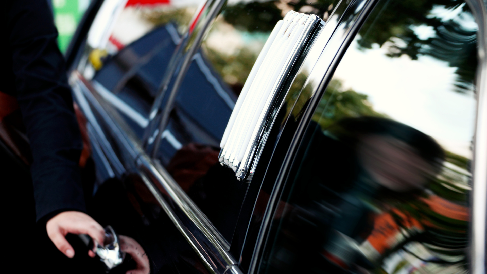 Person opening a black car door near a curb, with reflections on the glossy window.