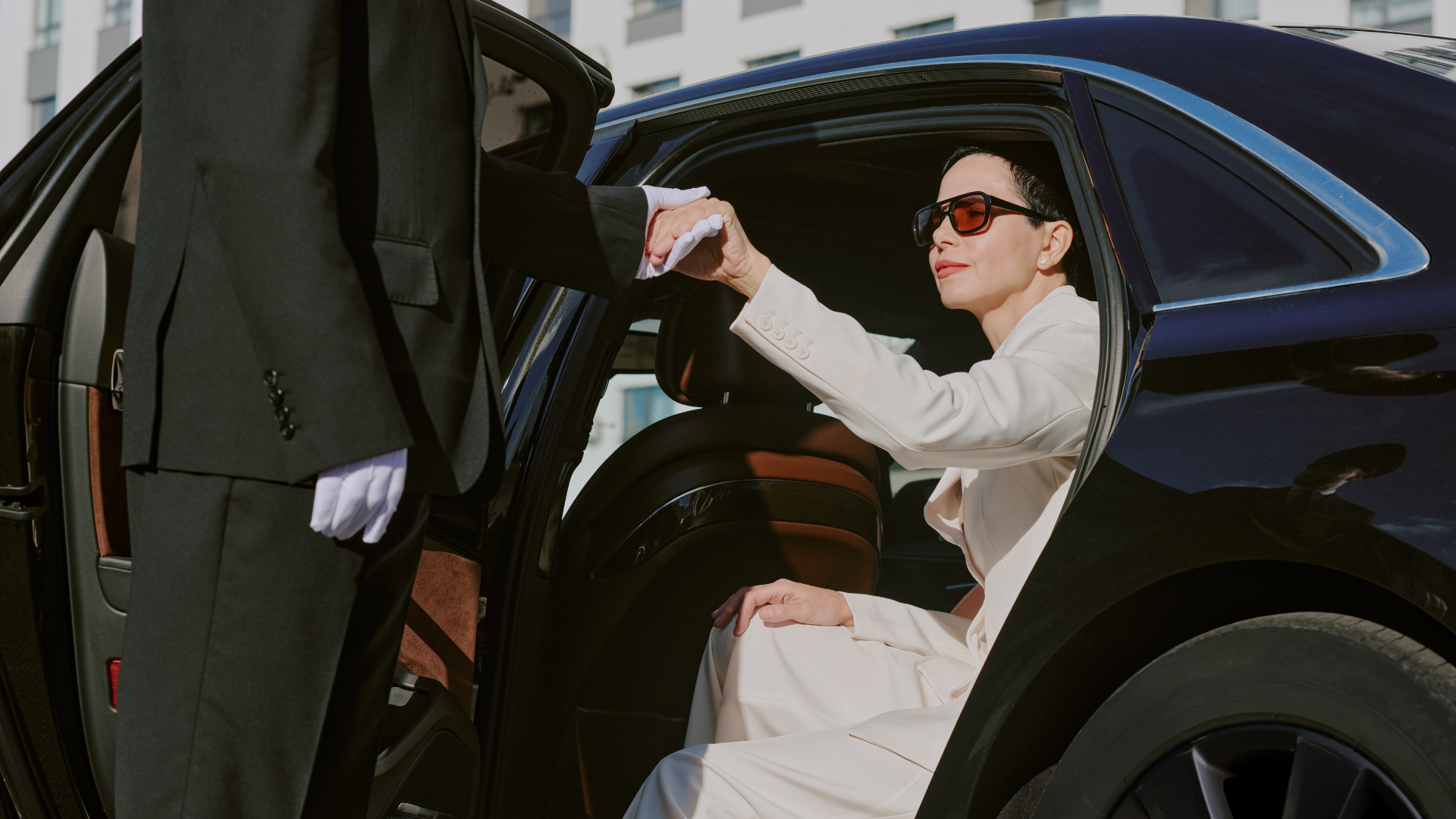 Woman in white coat and sunglasses stepping out of a black car, reaching toward the open door.