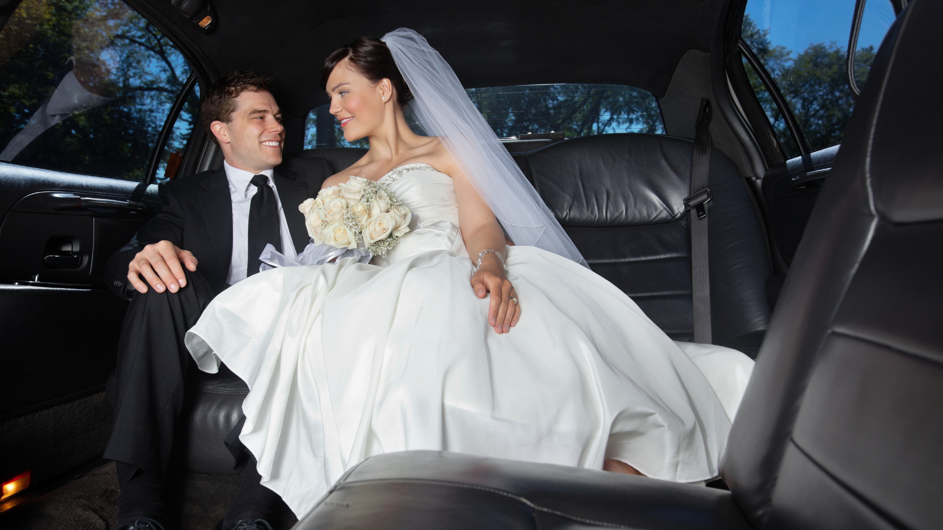 Bride and groom seated in a car, smiling at each other, with the bride in a white gown and veil.