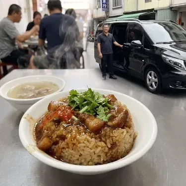 清水老街美食方形圖片。近距離捕捉清水阿財米糕(午餐建議)。糯米Q彈有勁,淋上肥而不膩、慢火細熬的滷肉汁,配上特製辣醬,畫面冒著煙火氣。背景略為模糊,可見包車司機(無帽、休閒服飾)在巷口黑色 Mercedes-Benz Vito 車旁等候,完美展現冷氣專車無縫接軌、免除曝曬之苦的包車價值。
