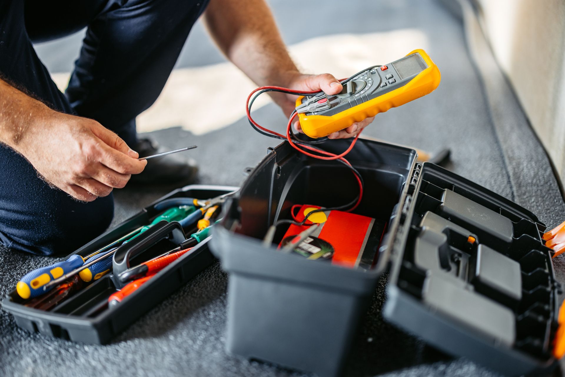 An electrician using a multimeter and screwdriver next to an open toolbox filled with tools.