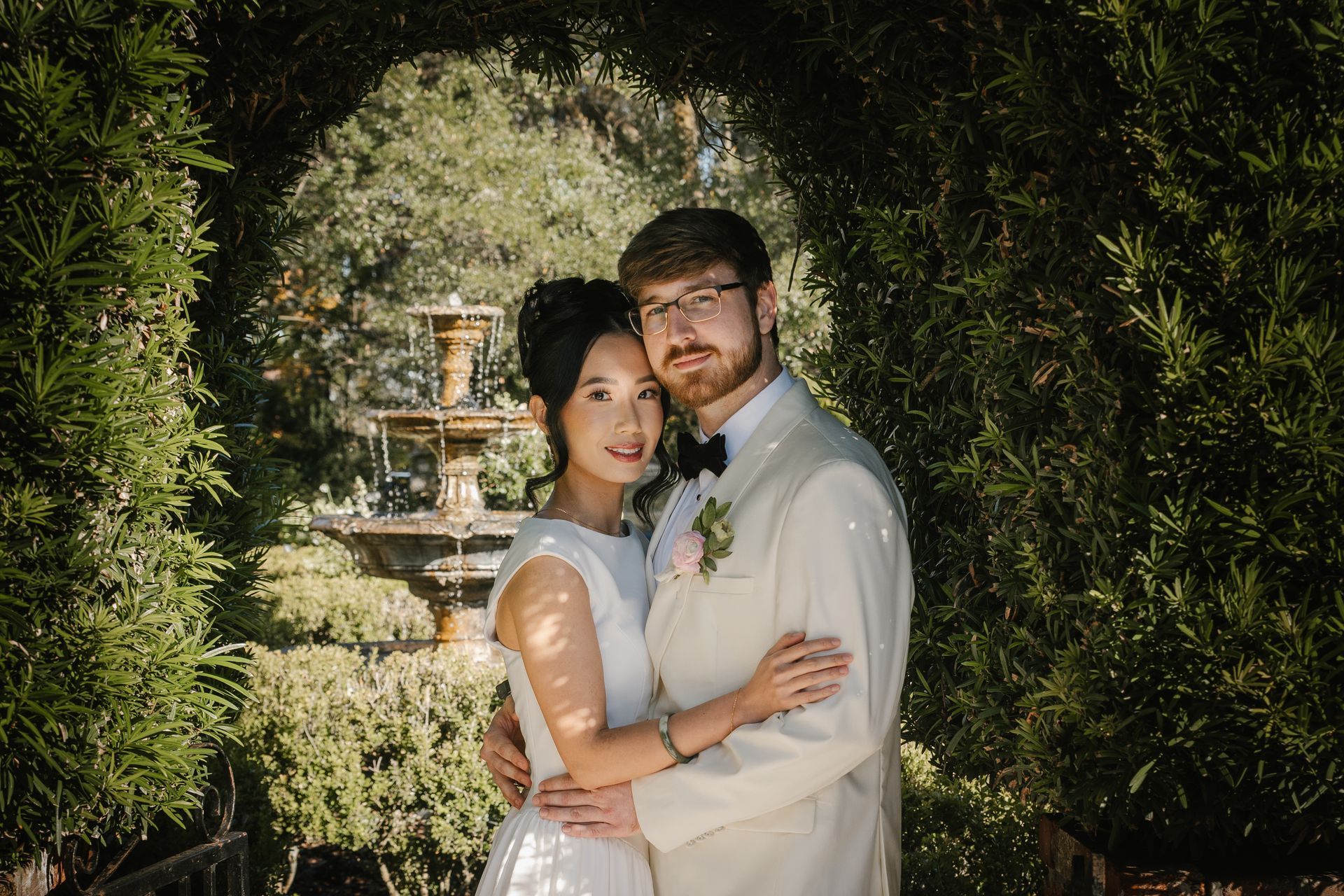 Bride in white dress holding groom under podocarpus hedge arch