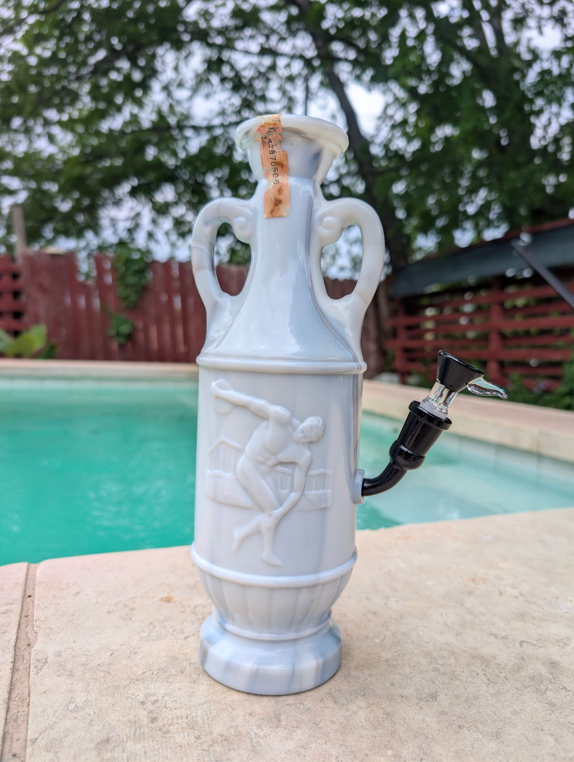 White decorative outdoor faucet ornament on a poolside patio, with a hose attached and turquoise pool in the background