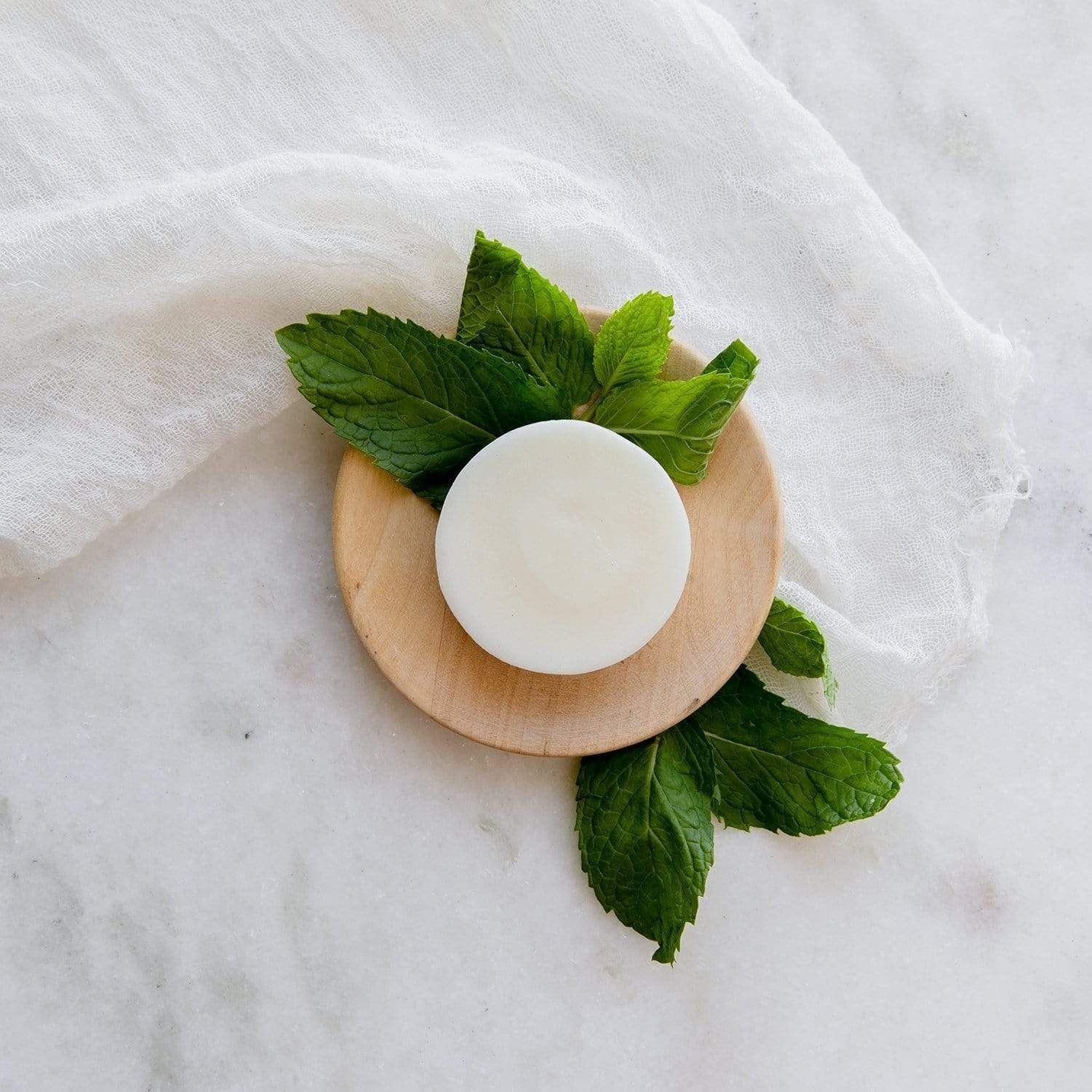 A white circular soap bar sits on a small wooden coaster, surrounded by fresh mint leaves on a white marble surface.