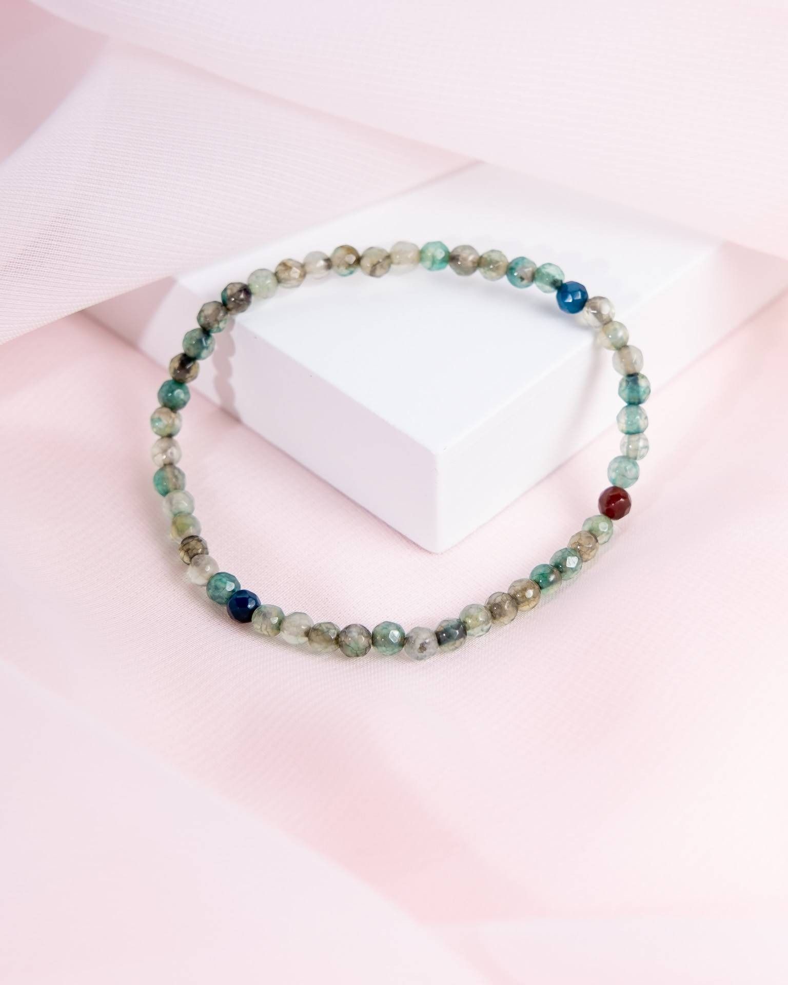 A beaded bracelet made of small, faceted green stones accented with blue and red beads, resting on a white display block.