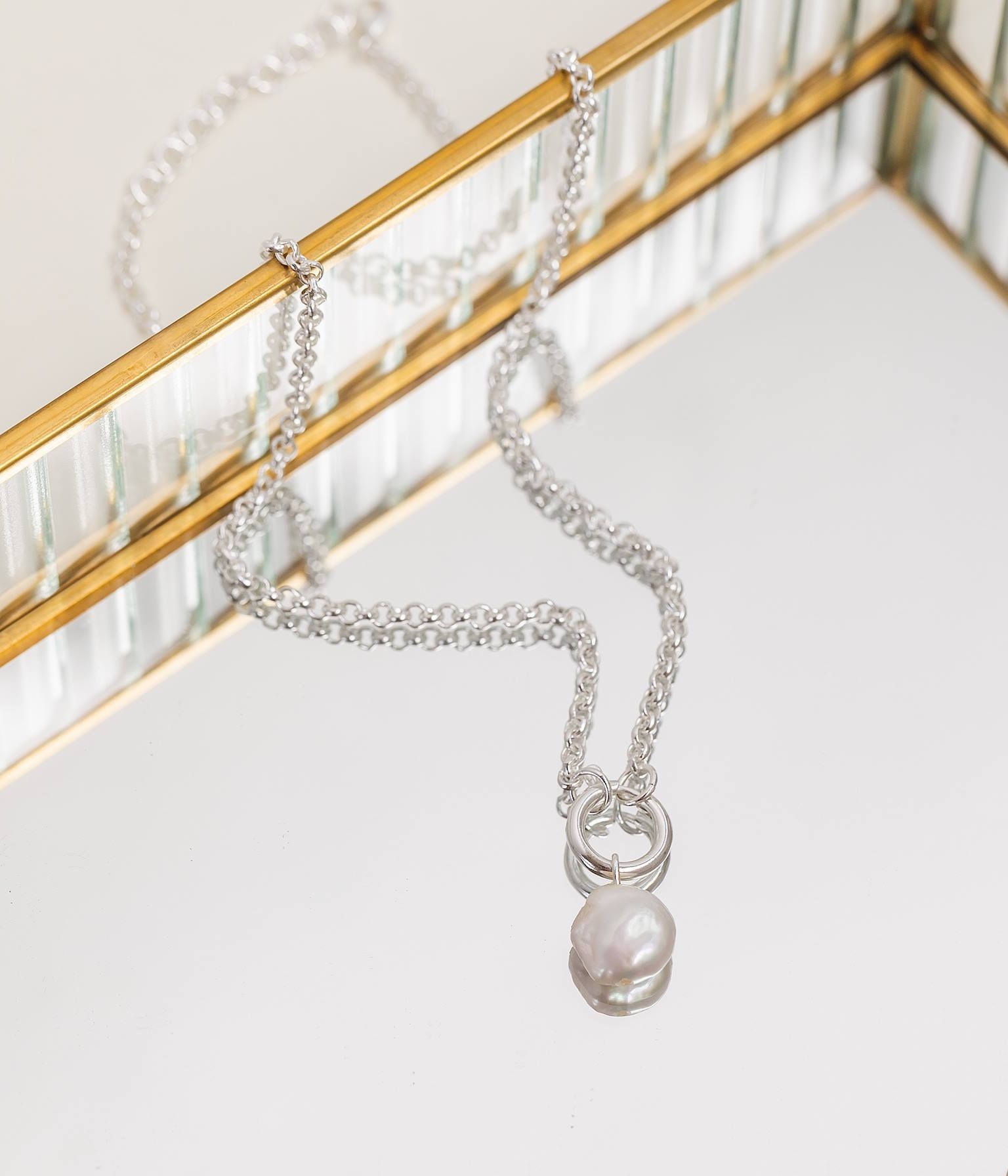 A silver chain necklace with a single round pearl pendant resting on a reflective surface against a gold-trimmed frame.