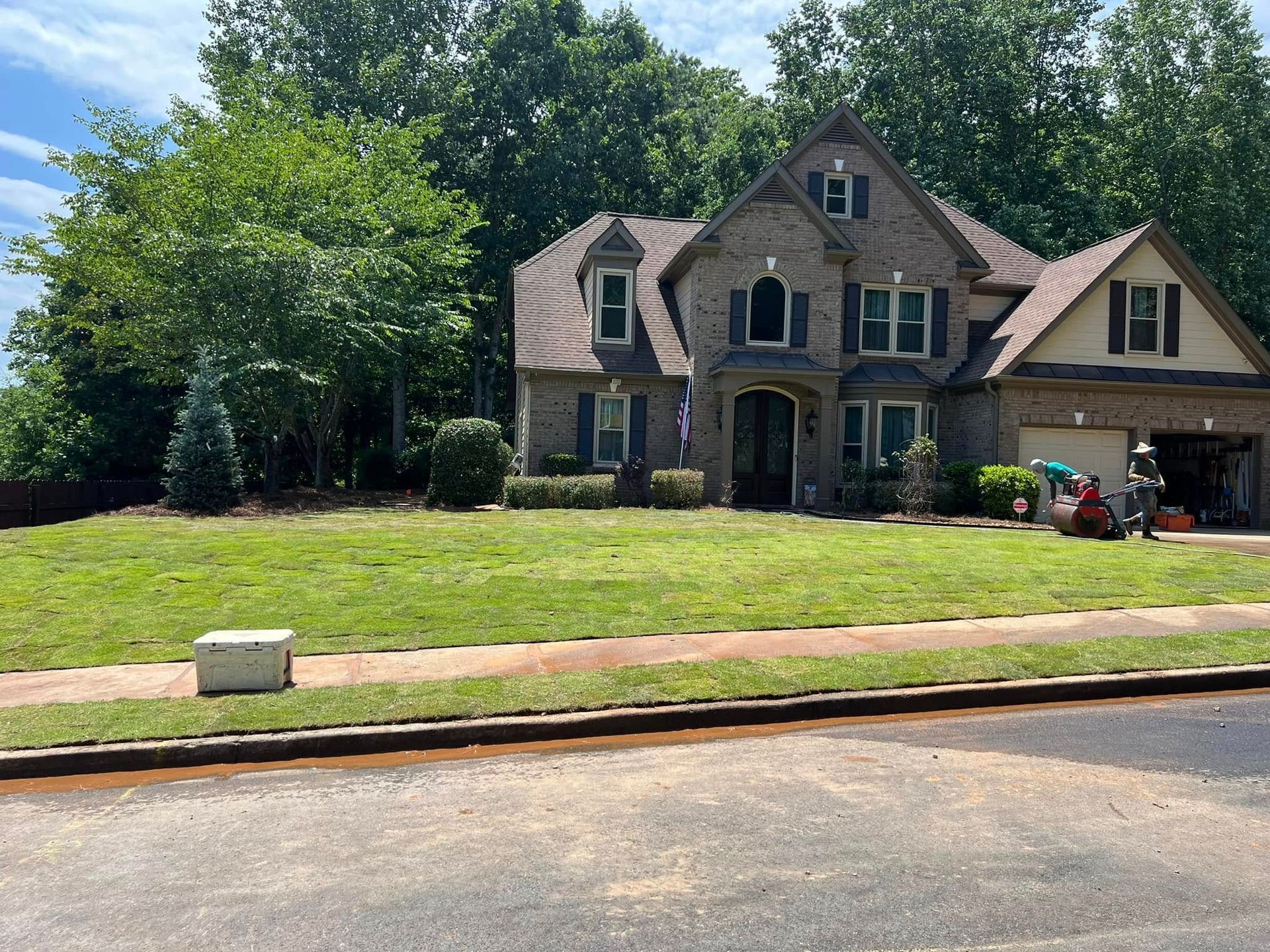 Large stone house with a front lawn, trees, and a driveway under a blue sky