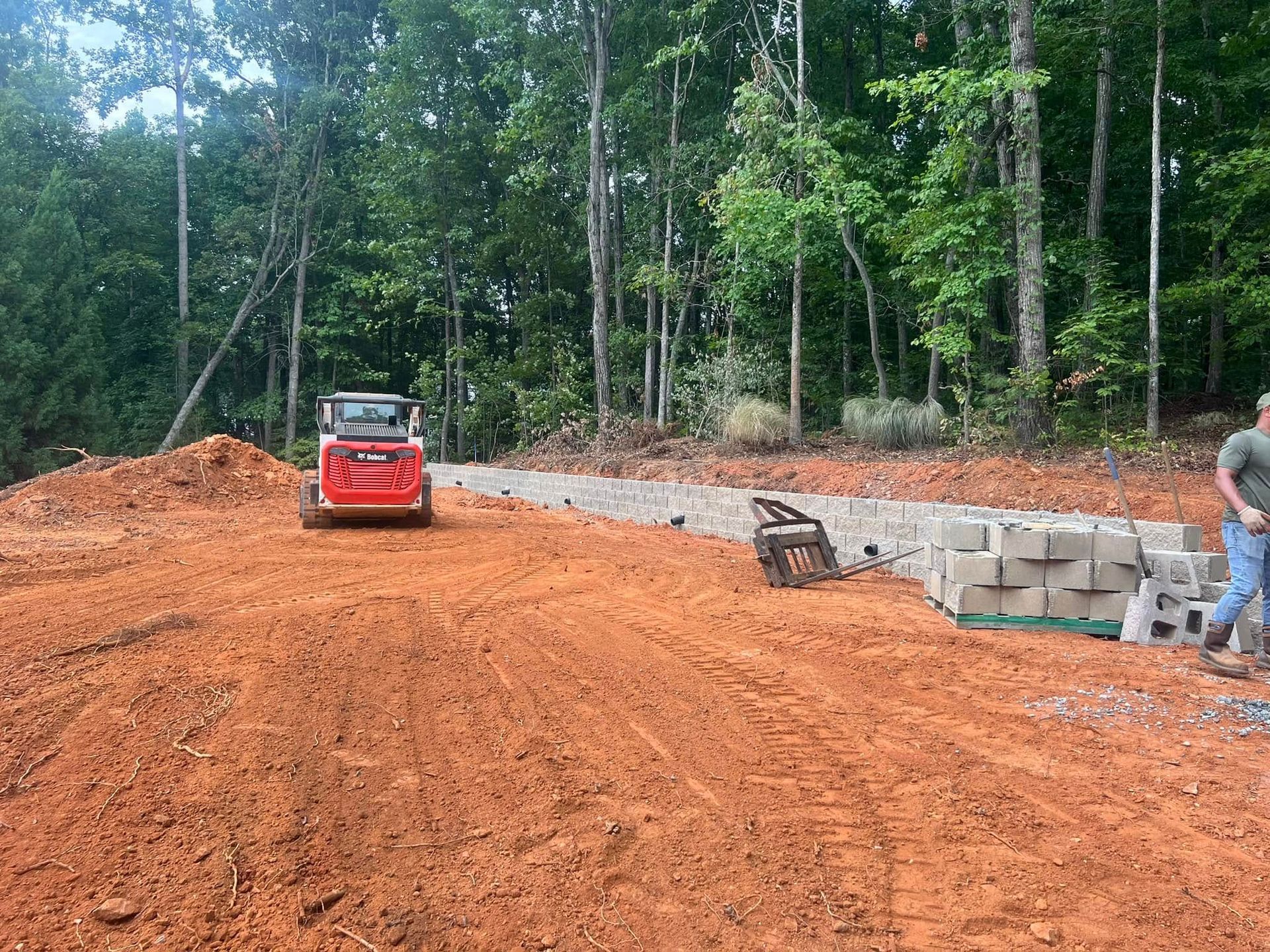 Red construction vehicle on a dirt driveway beside stacked cinder blocks, with workers near a wooded site.