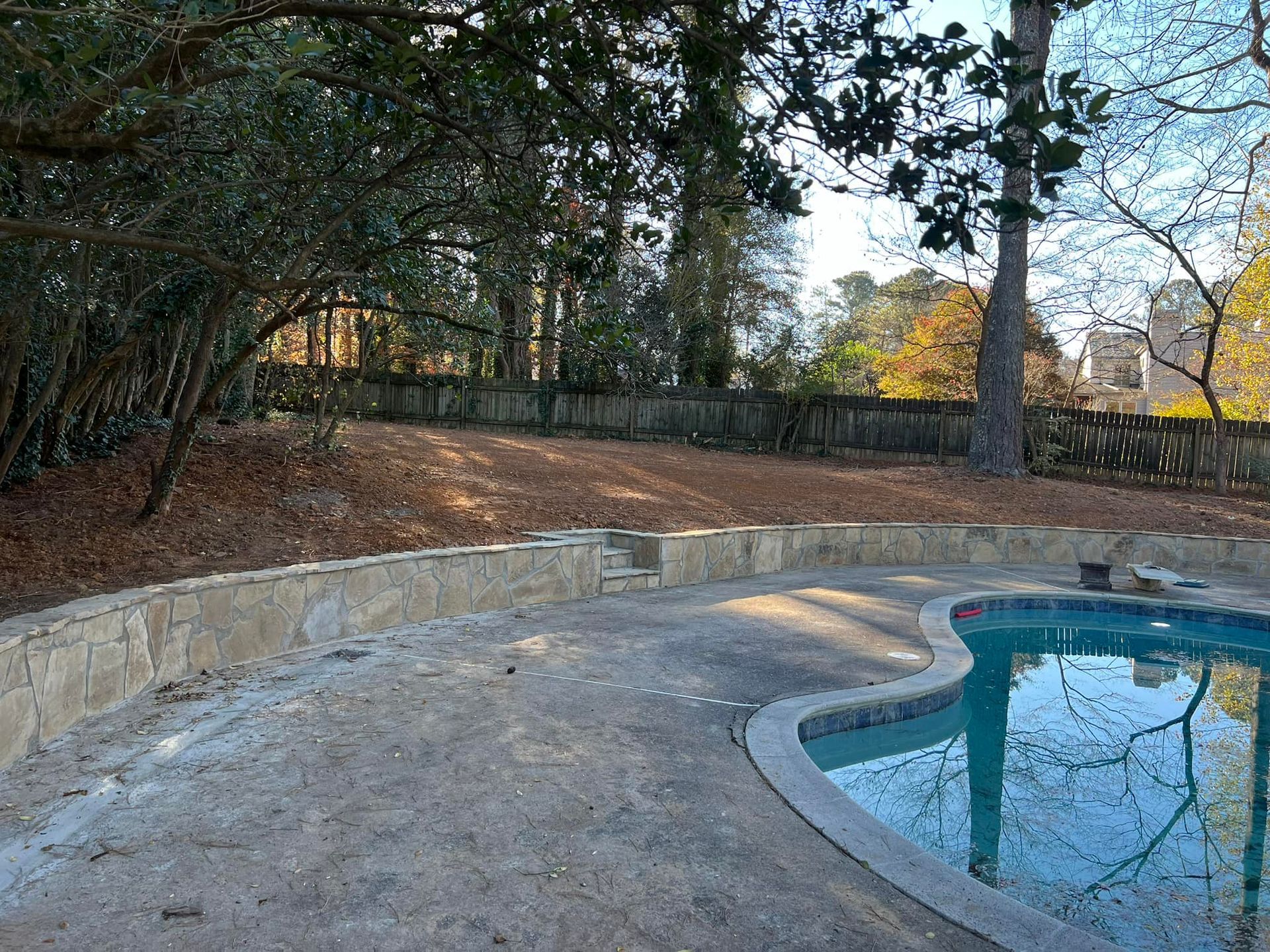 Curved stone pool edge beside trees and a fence in a wooded backyard yard