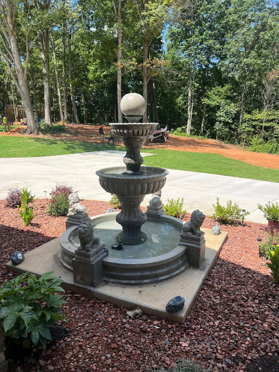 Tiered outdoor fountain on a circular stone base in a landscaped garden with red mulch and trees.