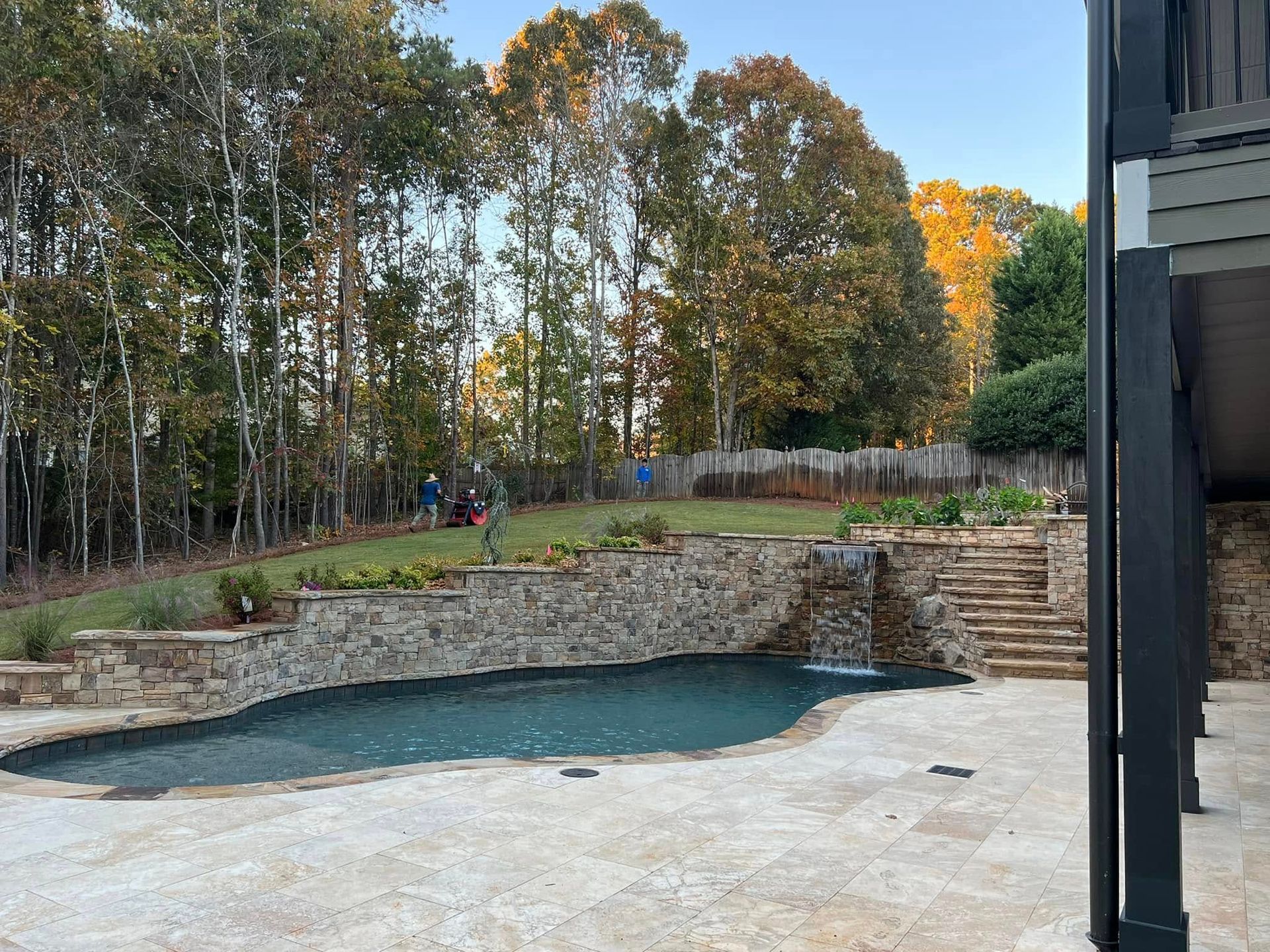 Backyard with an empty in-ground pool, stone retaining wall, and autumn trees beyond the patio