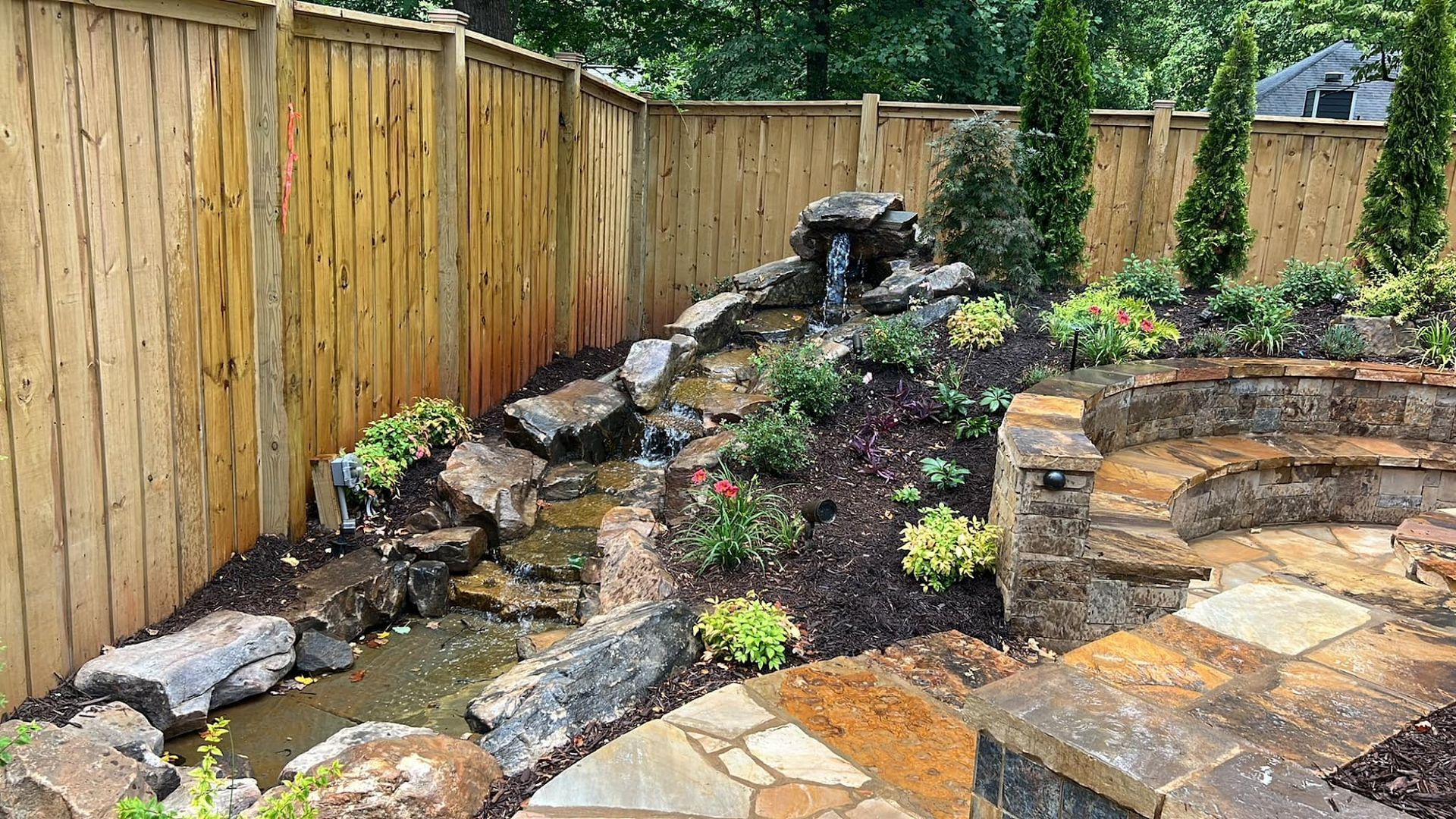 Backyard rock garden with a small waterfall, wooden fence, and stone patio under construction