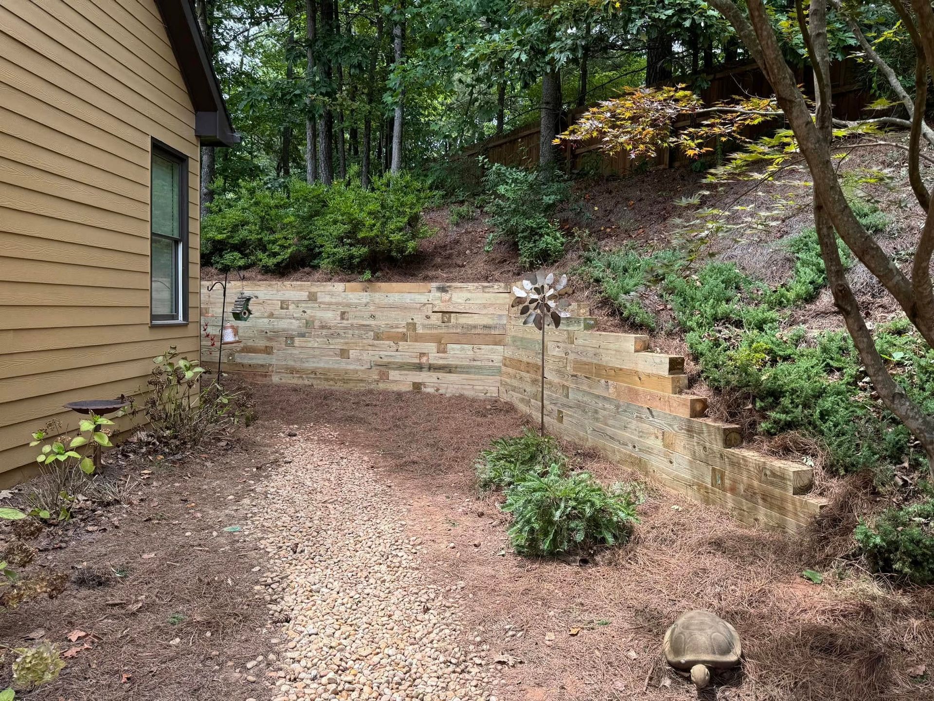 Small wooded backyard with a tan house wall, terraced retaining wall, and leaf-covered ground.