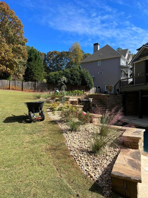 Sunny backyard with grass lawn, flower bed, and stone retaining wall beside a house